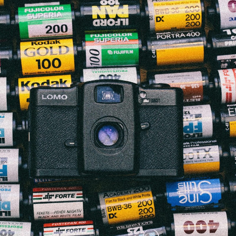 Чорно-біла плівкова камера LOMO на фоні касет Fujifilm, Kodak, Ilford та інших брендів.